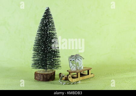Una miniatura in legno portante della slitta in un piccolo regalo accanto ad un luminoso verde albero di natale su uno sfondo verde chiaro Foto Stock