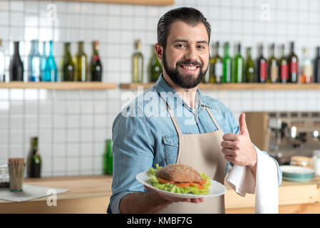 Entusiastico barbuto cameriere sentimento buono tenendo una piastra con il cibo Foto Stock