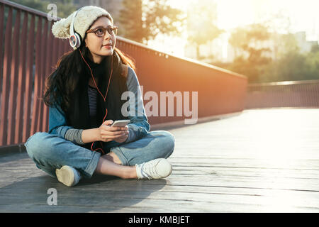Affascinante ed elegante asia donna gode di musica dello smartphone sul ponte della città, ragazza gode di giornata di sole nella stagione invernale. Foto Stock