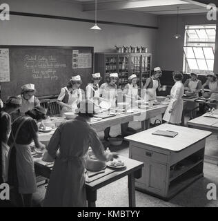 Degli anni Cinquanta, storico, cucina pratica classe, immagine mostra un gruppo di studenti di sesso femminile nella classe con insegnante imparando a cucinare un corso di due senza carne o pasto vegetariano, formaggio, uova e la torta di patate e un rhubard sbriciolate il budino, Inghilterra, Regno Unito. Foto Stock