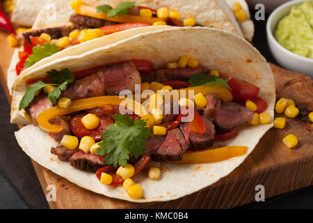 Tre Mexican tacos con carne di manzo in marmo, Black Angus e verdure su tavola di legno su una pietra scura sullo sfondo. Piatto messicano con salse guacamole e s Foto Stock