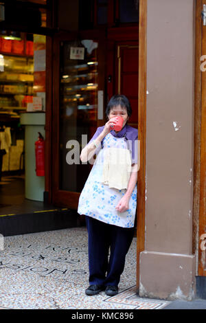 Vecchia Signora cinese in piedi nella porta del ristorante che indossa e grembiule e di bere da un bicchiere di plastica in Chinatown, Soho, London, England, Regno Unito Foto Stock