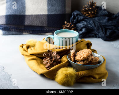 La tazza di tè con latte e biscotti con scaglie di cioccolato in un tenue giallo inverno sciarpa su sfondo grigio. pausa caffè per casa accogliente potrete rilassarvi con plaid Foto Stock
