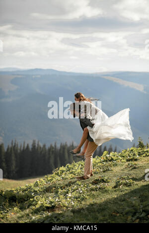 Sposi novelli stand, abbraccio, ridere e divertirsi durante il viaggio di nozze nei Carpazi. lo sposo porta sposa sulla sua schiena. Foto Stock