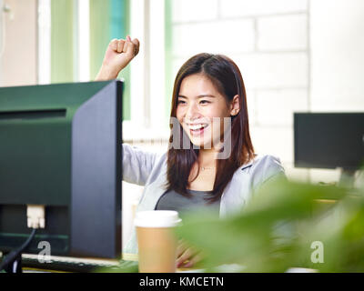 Giovani asian business donna che lavorano in ufficio utilizzando il computer desktop, felice ed entusiasta di buone notizie. Foto Stock