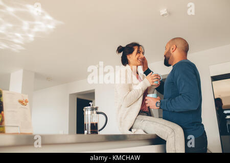 Coppia giovane avente caffè insieme a casa in cucina. Donna seduta sul banco di cucina con uomo in piedi da. Foto Stock