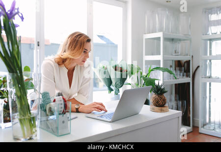 Fiorista femmina utilizzando laptop a contatore nel negozio di fiori. Piccolo negozio di fiori proprietario lavora su computer portatile nel suo negozio. Foto Stock