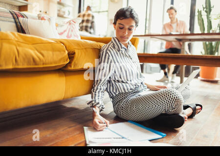Freelance femminile che lavora su un computer portatile, scrivendo su documenti cartacei in soggiorno Foto Stock