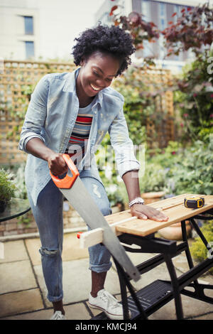 Donna sorridente con sega che taglia legno sul patio Foto Stock