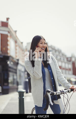 Sorridente giovane donna che si spostava in bicicletta, parlando al telefono cellulare sulla soleggiata strada urbana Foto Stock