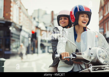 Sorridendo giovani donne amici che indossano caschi e scooter da guida sulla strada urbana Foto Stock