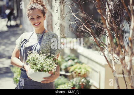 Ritratto sorridente fiorista femminile che tiene in vaso pianta al soleggiato magazzino Foto Stock