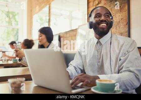 Un giovane sorridente con le cuffie che usano il computer portatile e bevono caffè al tavolo del caffè Foto Stock
