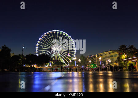 Place Masséna, Nizza, Francia, 5 dicembre 2017. Una bellissima giornata di sole con il cielo limpido nel bel mercatino di Natale si conclude con giochi di luce e di riflessi nell'acqua spruzzata piano delle fontane di Place Masséna, Nizza la storica piazza centrale al tramonto. La rilassata atmosfera del mercato, con cibo e dono bancarelle, bambini giostre, luci colorate e una gigantesca ruota panoramica Ferris splendidamente cattura lo spirito della Cote d Azur stagione festiva. Foto Stock