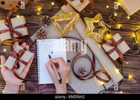 Vista superiore mani femminili iscritto lista regali al notebook per un amato popolo per natale sul tavolo di legno con decorazioni, luci garland, notebook, craft p Foto Stock