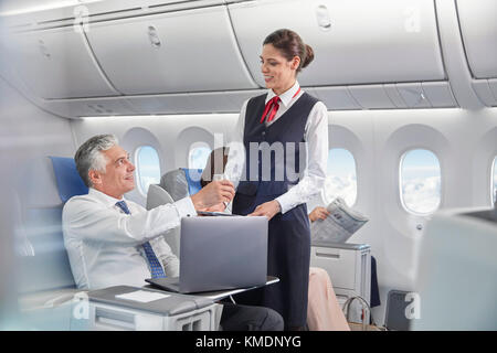 Assistente di volo che serve drink a un uomo d'affari che lavora su un computer portatile aereo Foto Stock