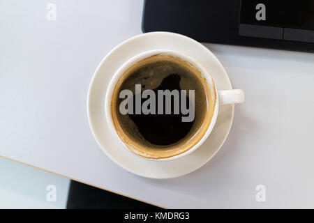 Una tazza di caffè nero in una tazza bianca su una scrivania in ufficio accanto a un nero tastiera portatile Foto Stock