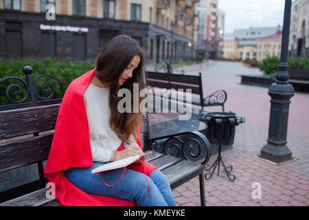 Ragazza attraente con i capelli lunghi siede su una panca, coperta in una coperta rossa, in un nuovo quartiere residenziale e scrive i suoi pensieri in un taccuino rosso. Foto Stock