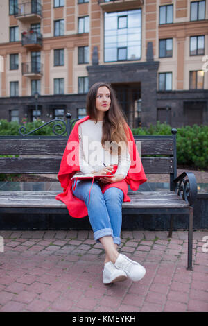 Ragazza attraente con i capelli lunghi siede su una panca, coperta in una coperta rossa, in un nuovo quartiere residenziale e scrive i suoi pensieri in un taccuino rosso. Foto Stock