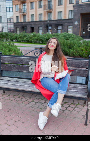 Una ragazza attraente siede su una panca e scrive i suoi pensieri sullo sfondo urbano in un taccuino rosso. Indossa un maglione bianco, jeans blu e un rosso Foto Stock