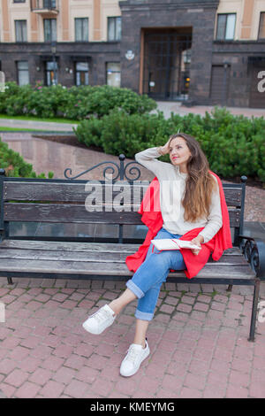 Una ragazza attraente siede su una panca e scrive i suoi pensieri sullo sfondo urbano in un taccuino rosso. Indossa un maglione bianco, jeans blu e un rosso Foto Stock