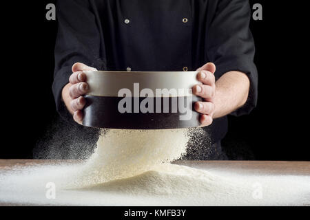 Mani maschio setacciare la farina dal vecchio crivello su una vecchia cucina in legno tavolo. isolato su sfondo nero. Foto Stock
