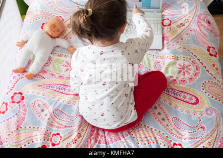 Poco ragazza seduta nel letto e giocare online con il computer portatile. Ella sta toccando lo schermo Foto Stock