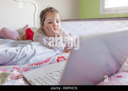 Bambina che giace nel letto e guardare i cartoni animati con un computer portatile nella sua camera da letto. Lei sembra divertirsi Foto Stock