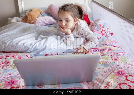 Bambina che giace nel letto e guardare i cartoni animati con un computer portatile nella sua camera da letto. Lei sembra divertirsi Foto Stock