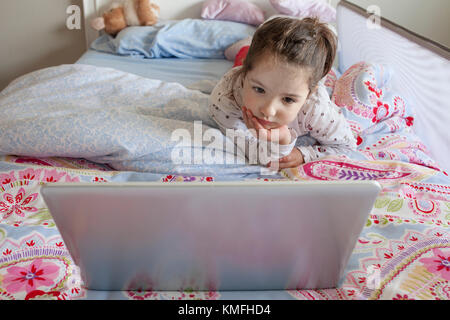 Bambina giacente in letto e giocare con un computer portatile nella sua camera da letto. Guarda attento Foto Stock