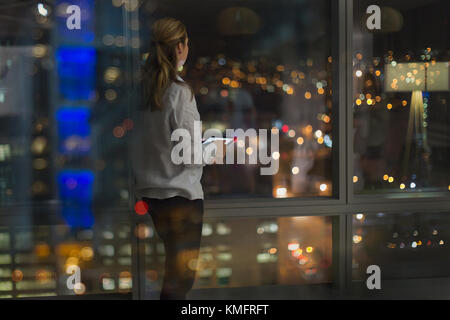 Pensiva donna d'affari con tablet digitale che lavora tardi, guardando fuori dalla finestra dell'ufficio urbano di notte Foto Stock