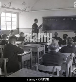 Degli anni Cinquanta, storica aula scolastica, insegnante maschio in abito tradizionale a blackboard insegnamento scuola ragazzi seduti a scrivanie singole calcoli, Inghilterra, Regno Unito. Foto Stock