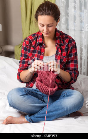 Close-up di una donna in stato di gravidanza in un plaid shirt e jeans seduti su un letto e una maglia con aghi da maglia da una naturale rosa stringa di lana di sudore Foto Stock