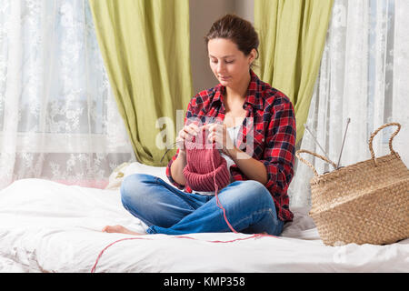 Dai capelli scuri giovane donna incinta in plaid shirt e jeans maglieria con aghi da maglia da naturale rosa filo di lana maglione su letto di casa Foto Stock