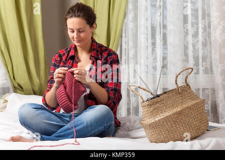 Una donna in stato di gravidanza in un plaid shirt jeans e si siede sul letto e maglieria con aghi da maglia da una naturale rosa stringa di lana di un maglione, in backg Foto Stock