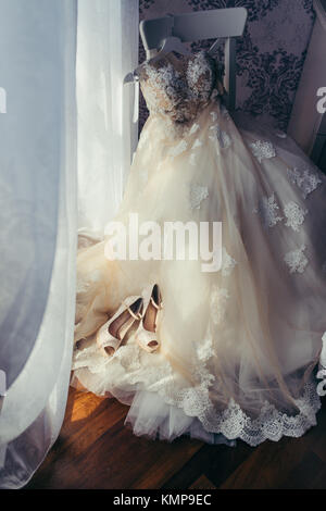 La visione verticale del bianco elegante lungo abito da sposa con fiori appesi sulla sedia. il beige tacchi alti sono collocati sul vestito. Foto Stock