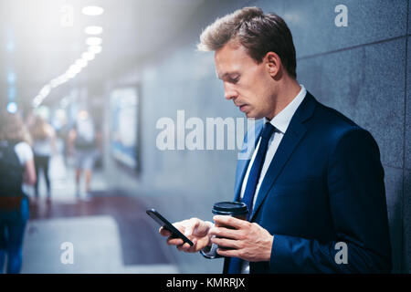 Giovane imprenditore a bere caffè e la lettura dei messaggi di testo sul suo cellulare mentre in piedi su una piattaforma della metropolitana durante i suoi spostamenti di mattina Foto Stock