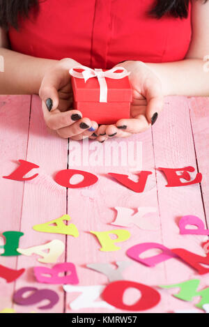 Donna mani tenendo una confezione regalo rossa su una rosa tavolo in legno e piena di carta multicolore di lettere e la parola amore scritto fuori di essi. Foto Stock