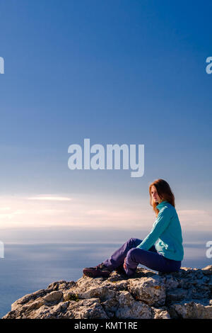 Sporty giovane donna seduta in una sommità rocciosa della montagna contro il blu del cielo e del mare. Di fronte alla telecamera Foto Stock