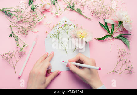 Mani femminili di scrivere con la matita sulla busta aperta con disposizione dei fiori . Fioraio attrezzature di decorazione su rosa sfondo tabella, vista dall'alto. Invitati Foto Stock