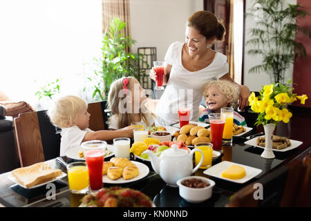 Famiglia sana colazione a casa. Madre e bambini a mangiare la frutta tropicale, pane tostato, formaggio e salsicce. Bambini bere freschi succo pressato sul soleggiato Foto Stock