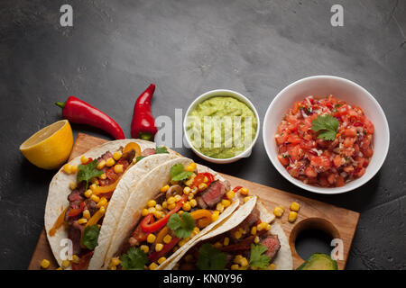 Tre Mexican tacos con carne di manzo in marmo, Black Angus e verdure su tavola di legno su una pietra scura sullo sfondo. Piatto messicano con salse guacamole e s Foto Stock