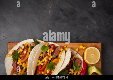 Tre Mexican tacos con carne di manzo in marmo, Black Angus e verdure su tavola di legno su una pietra scura sullo sfondo. Piatto messicano con salse guacamole e s Foto Stock