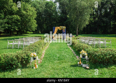 Le impostazioni di bella all'aperto per la cerimonia nuziale e la celebrazione. Splendidamente decorate arco con fiori e tante vuote con sedie in legno in luogo scenico Foto Stock