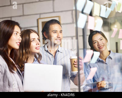 Team di giovani asiatici e caucasici imprenditori discutere di affari con computer portatile, tavoletta digitale e note adesive in ufficio. Foto Stock