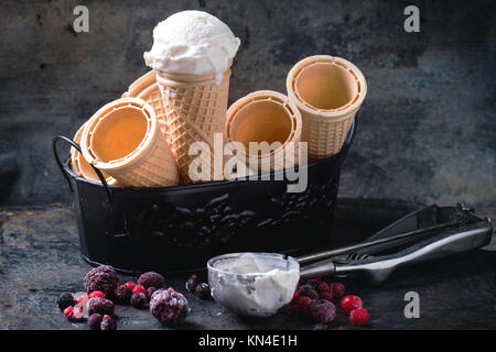 Gelato alla vaniglia in coni e svuotare i coni di waffer, servita nel recipiente di metallo con frutti di bosco surgelati e cucchiaio su tavola nera. Vedere serie Foto Stock