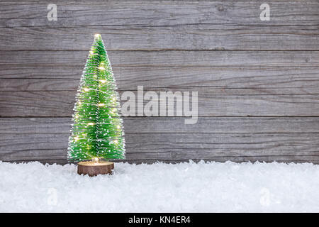 Piccola verde artificiale albero di Natale illuminato con luci delle vacanze sulla neve contro di legno sfondo rustico Foto Stock