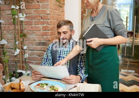 Ritratto di maturo uomo barbuto menu di lettura in moderno cafe con la giovane cameriera dandogli ordinazione di consulenza sul business lunch Foto Stock