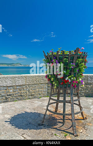 Un basket fuoco con fiori sul lungomare di Lyme Regis con cappuccio dorato al di là su Jurassic Coast Sito Patrimonio Mondiale, Dorset, England, Regno Unito Foto Stock