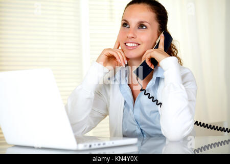 Bella giovane donna a conversare al telefono al posto di lavoro nella parte anteriore del computer portatile.